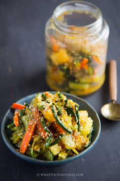 a bowl filled with vegetables next to a jar of pickles and spoon on a table