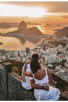 a man and woman sitting on top of a stone wall looking out over the city