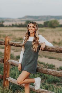 a woman leaning on a wooden fence posing for a photo in front of an open field