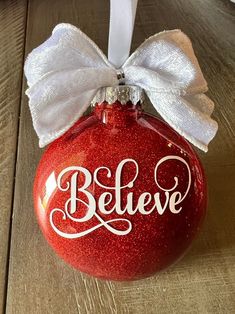 a red ornament with a white bow and believe written on the front, sitting on a wooden surface