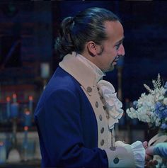 a man with long hair wearing a blue coat and white gloves holding a bouquet of flowers