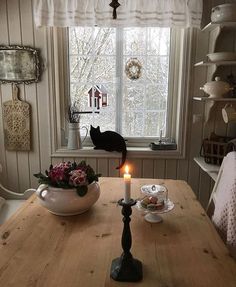 a table with a candle on it in front of a window and a cat looking out the window