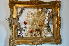 an ornate gold frame with dried flowers and leaves hanging on the wall in front of a white wall