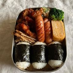 an assortment of sushi in a metal container on a white tablecloth with broccoli and bread