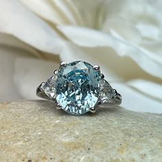 "The ring pictured is simulated diamond aquamarine #7126 -Approximate total carat weight: 5.00ctw diamond equivalent -Center Stone Size: 11x9mm - approx. 4.20ct diamond equivalent -Center Stone Shape: oval cut -Side Stones Size: 2 Trillions 5x5mm - approximately 0.80ctw diamond equivalent -Gem Type: simulated diamond -Stone Clarity: VVS1 -Stone Color: Blue Aquamarine -Moh's Scale: 8.5 hardness -Metal Type and Purity: 14k white gold -Setting: 4 prong tulip head -Stock Ring Size: 6 -Country of Man Trillion Engagement Ring, Engagement Ring Three Stone, Ring Three Stone, Blue Sapphire Necklace, Aquamarine Engagement Ring, White Gold Set, Oval Ring, Diamond Wedding Ring, Oval Rings