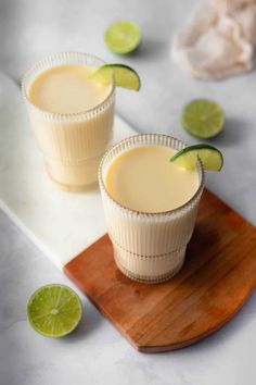 two glasses filled with drinks sitting on top of a cutting board next to limes