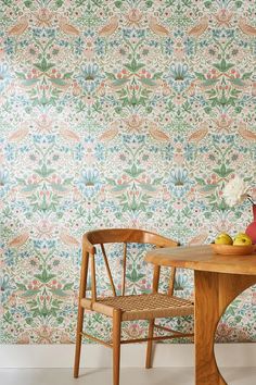 a wooden chair sitting next to a table with fruit on it in front of a floral wallpaper