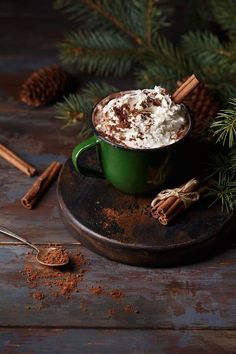 a cup of hot chocolate with whipped cream and cinnamon sticks on a wooden table next to pine cones