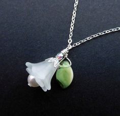 "Fresh as a bright Spring day, is this white lucite flower pendant necklace with it's jaunty leaf, and Silver-plated findings.  Frilly layered flowers with a White Swarovski pearl peeping out, and green glass leaf wire-wrapped beside it, are hung from a delicate Silver-plated chain. Two frosted white lucite ruffled lily flowers are layered together with the 6mm pearl.  The flowers are capped with a pointed 'petal' bead cap, and a 3mm Czech fire-polished crystal in Aurora Borealis. A small green White Sterling Silver Charm Necklaces With Flower Charm, White Sterling Silver Charm Necklace With Flower, Delicate White Flower Charm Necklaces, Delicate White Flower Charm Necklace, Delicate Green Sterling Silver Necklaces, Green Flower Pendant Jewelry For Wedding, Delicate Green Sterling Silver Necklace, Green Sterling Silver Necklace With Flower Pendant, Elegant Green Sterling Silver Charm Necklace