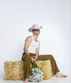 a man wearing a cowboy hat sitting on hay bales with flowers in his lap