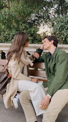 a young man and woman sitting on a bench talking to each other in front of some trees