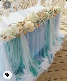 the table is set up on the beach for an ocean themed wedding ceremony with white flowers and blue tulle