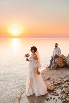 the bride and groom are sitting on rocks by the water at sunset, with the sun setting behind them