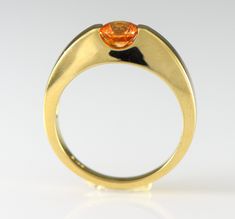 A brilliant orange gem Spessartite Garnet is bar set in a streamlined stylish 14k yellow gold ring. Spessartite, a rare form of garnet, also called mandarin or Malaya garnet, ranges from a yellow-orange through brilliant orange to orange-red. They combine very good hardness with excellent brilliance and sparkle. Spessartite is found in Africa and like all garnets is untreated. Garnet is the birthstone for January. Metal: 14k Yellow Gold The spessartite garnet is a 5.5mm round and weighs .84 cara Modern Yellow Gold Sapphire Ring With Polished Finish, Modern Orange Oval Rings, Fine Jewelry Orange Sapphire Anniversary Ring, Modern Topaz Ring With Center Stone In Yellow Gold, Classic Orange Rings With Polished Finish, Modern Orange Gemstone Rings, Modern Yellow Gold Topaz Ring With Center Stone, Modern Sapphire Ring With Polished Finish And Round Band, Orange Sapphire Ring For Anniversary