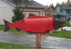 a red fish mailbox sitting on top of a wooden post in front of a house