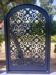 an iron gate with a tree on it in the middle of a park area, surrounded by trees and bushes