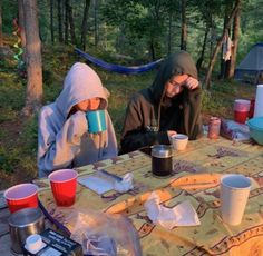 two people sitting at a picnic table with cups in front of them and camping gear on the ground