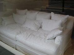 a white couch with lots of pillows on it's back and sides, sitting in front of a window