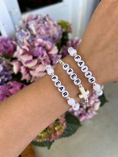 two bracelets with words on them and a cross in the middle, sitting next to flowers