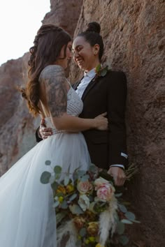 a bride and groom standing next to each other in front of a rock wall with their arms around each other