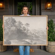 a woman holding up a large painting in front of a house with potted plants