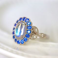 Mesmerizing Blue Moonstone Halo Ring. Moonstone engagement ring. Blue sapphires & diamonds. Edwardian Moonstone. 14K White Gold. US 7.25 A beautiful and unique moonstone ring, created with precision cut sapphires in 14K white gold, a luxurious and high-quality piece of jewelry, perfect to express your love. 💎 PREMIUM RING -- A brand new moonstone ring, personally designed and made with the highest quality of 14K white gold.  ✨ CLASSIC BRILLIANCE -- Designed with a beautiful blue moonstone at th Blue Round Sapphire Ring With Rose Cut Diamonds, Elegant Sapphire Moonstone Birthstone Ring, Elegant Sapphire Moonstone Ring In Oval Shape, Elegant Sapphire Color Moonstone Ring, Anniversary Sapphire Gemstones With Halo Setting, Sapphire Moonstone Ring Fine Jewelry Gift, Moonstone Ring With Diamond Halo Setting, Elegant Sapphire Moonstone Ring For Anniversary, Round Sapphire Jewelry With Rose Cut Diamonds