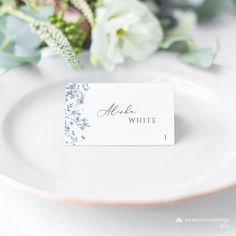 a place card sitting on top of a white plate with flowers in the back ground