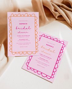 two pink and orange wedding cards on top of a white tablecloth next to a brown cloth