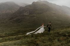 a bride and groom holding hands while walking through the grass with mountains in the background