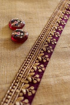 Wine and gold - a colour combination that spells royalty. This elegant trim displays a row of little flowers in gold. It is created with gold zari embroidery on a wine cotton silk fabric. Embellished with tiny sequins. Regal and unique, will add a dash of glamour to anything it touches. Use it to embellish your denim jackets, or border your sarees. Or create home décor like lamps or curtain ties. Or accessories like hair bands. One trim, countless possibilities. Price for 1 mtr Size - 3.5 cm W N Cotton Embroidery Sarees, Gold Silk Thread Wedding Sets, Purple Embroidered Fabric With Zari Work For Navratri, Purple Zari Work Embroidered Fabric For Navratri, Gold Embroidered Raw Silk Sets, Navratri Purple Zari Work Embroidered Fabric, Eid Purple Blouse Piece With Embroidered Border, Gold Zari Work Sets For Festivals, Traditional Gold Sets With Zari Work