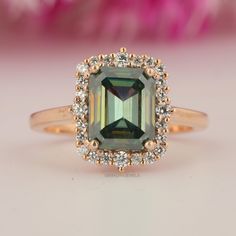 an emerald colored stone surrounded by white and yellow diamonds on a gold ring with pink flowers in the background