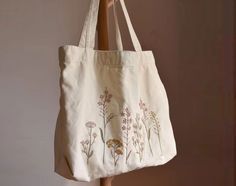 a white tote bag with flowers on it hanging from a wooden pole in front of a wall