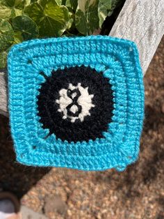 a blue crocheted square with a black and white design on the center is hanging from a wooden post