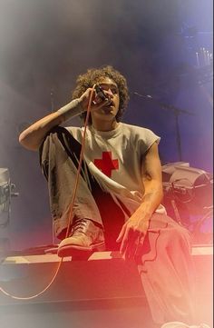 a man sitting on top of a stage while holding a microphone to his ear and wearing a red cross t - shirt