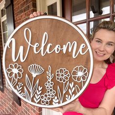 a woman holding up a wooden sign that says welcome with flowers and leaves on it