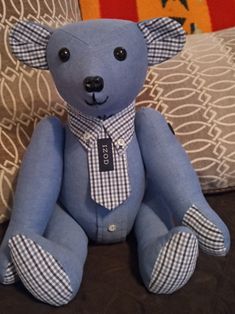 a blue teddy bear wearing a tie and checkered shirt sitting on a couch with pillows in the background