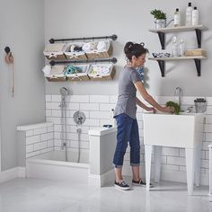 a woman standing at a sink in a white tiled bathroom with shelving and shelves