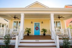 a blue door sits on the front porch of a yellow house with white railings and potted plants