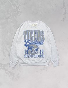 **Officially licensed with the University of Memphis** Show the love for your favorite team in our one and only Memphis Tigers Football oversized 90's sweatshirt. - Features Tigers Football with a helmet featuring the official Tigers logo with 1912 beside it and The University of Memphis, Established 1912. Memphis Tennessee underneath all in a blue, gray and white ink - Heat print transfer that is heat pressed onto each sweatshirt- Printed on a soft, 90's style oversized crewneck sweatshirt- Cot Collegiate Sweatshirt For Football Season, Collegiate Sweatshirt For College Football Season, Collegiate Sweatshirt With Team Logo For Football Season, Collegiate Style Sweatshirt With Team Logo For Football Season, Collegiate Sweatshirt With Team Logo, College Football Season Fan Apparel Sweatshirt, Collegiate Style Sweatshirt For Sports Events, Throwback Crew Neck Top For Game Day, College Team Logo Hoodie With Crew Neck