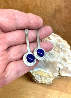 10mm Lapis Lazuli cabochons are bezel set on a larger Argentium Sterling silver disc, which is soldered to a hammered wire to make a beautiful dangly earring set.  Drilled holes at the tip of the hammered wires hold the ear wires.  The round silver disc is textured with hash marks around its perimeter.  They hang 2.5 inches with the ear wire being .75 inches of that. These beauties have definitely a feminine vibe with a modern feel. Argentium sterling silver is a recycled metal with more fine silver than regular sterling silver, but an added bit of germanium to help make it tarnish resistant.  All my metal and wire is Argentium 35. Lapis is noted for being a powerful stone for thinking and spirituality.  It is said to increase self-knowledge and awareness of your thoughts. Modern Sterling Silver Earrings With Cabochon, Round Metal Cabochon Earrings, Modern Cabochon Earrings For Gifts, Modern Cabochon Earrings Gift, Round Bezel Set Cabochons For Gifts, Hand Forged Blue Round Earrings, Blue Hammered Earrings For Gift, Sterling Silver Bezel Set Earrings, Sterling Silver Bezel Set Round Earrings