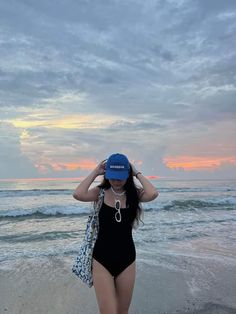 a woman in a black swimsuit and blue hat standing on the beach at sunset