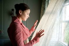 a woman standing in front of a window with her hands on the sheer curtain and looking out