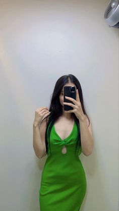 a woman in a green dress taking a selfie with her cell phone while standing against a wall