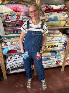 a woman in overalls standing next to a rack of quilts and other fabrics