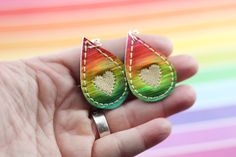 a hand holding a pair of colorful earrings in front of a rainbow colored background with white stitching