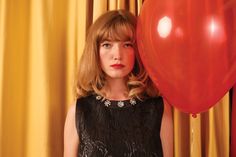 a woman standing next to a red balloon in front of a yellow curtain with gold drapes