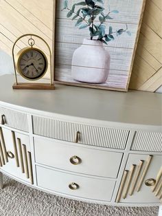 a white dresser with a clock on top of it next to a painting and vase