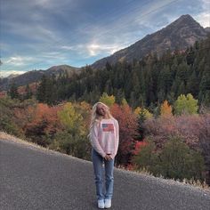 a woman standing on the side of a road in front of trees with fall colors