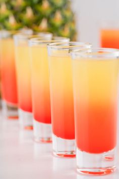 several glasses filled with different colored drinks on top of a white table next to pineapples