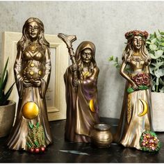 three statues of women holding pots and bowls on a table next to a potted plant